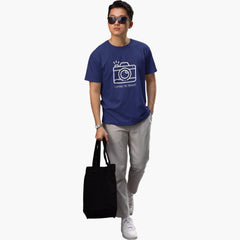 Person wearing a blue t-shirt with a camera graphic and text, holding a black tote bag on a white background