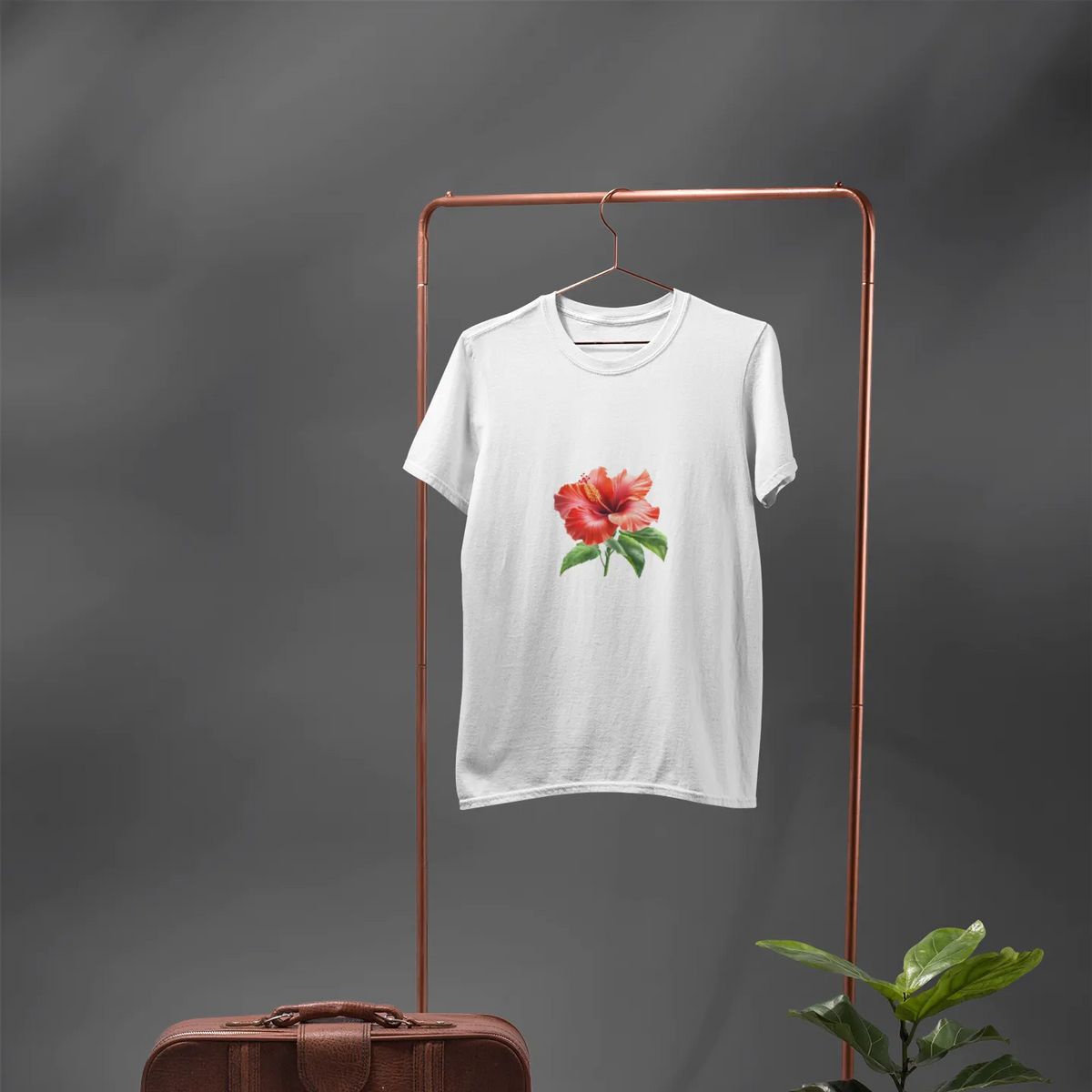 White t-shirt with a red flower design on a copper stand against a gray background, with a brown leather suitcase and potted plant.