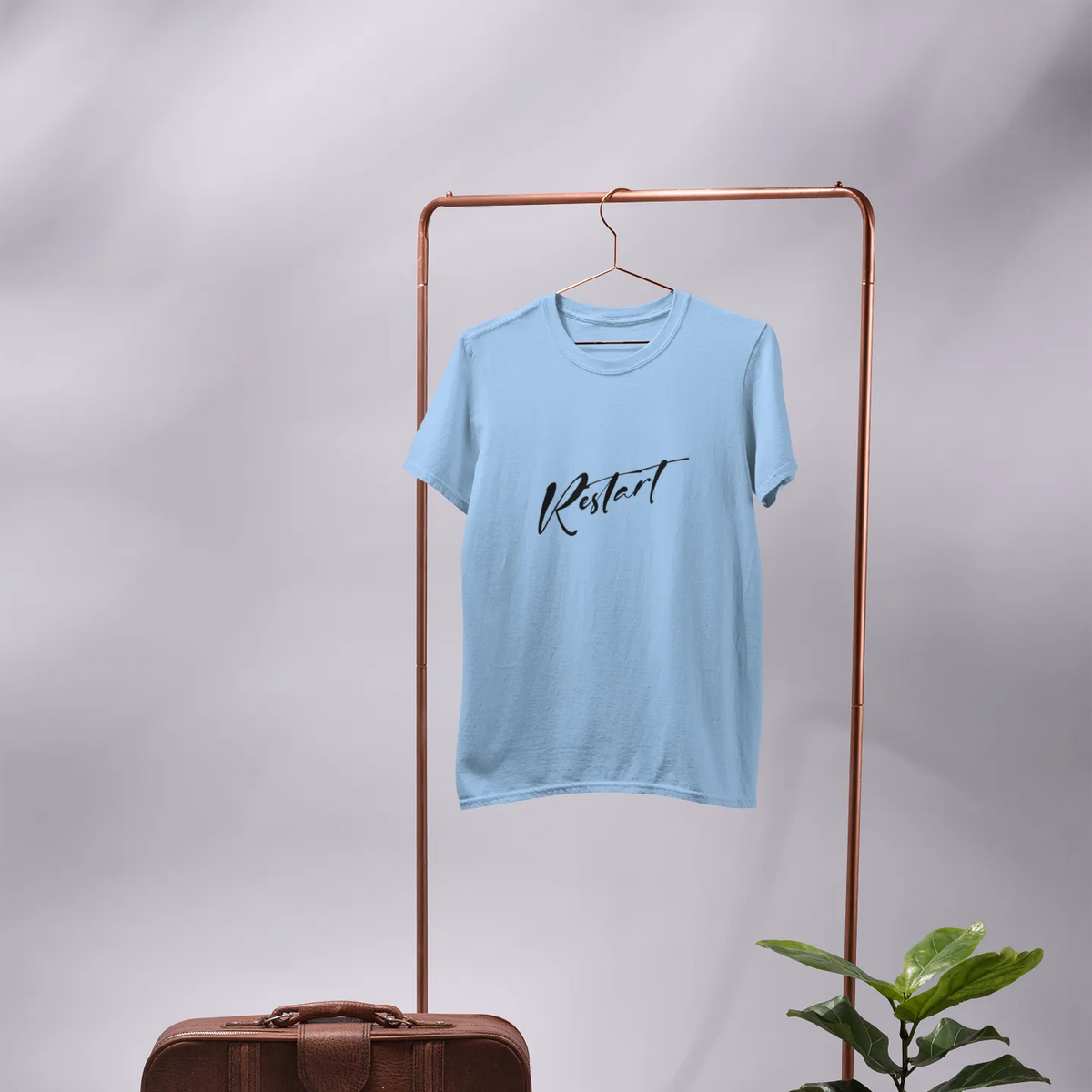 Light blue t-shirt with 'Kermit' text on a hanger, next to a brown suitcase and potted plant on a gray background