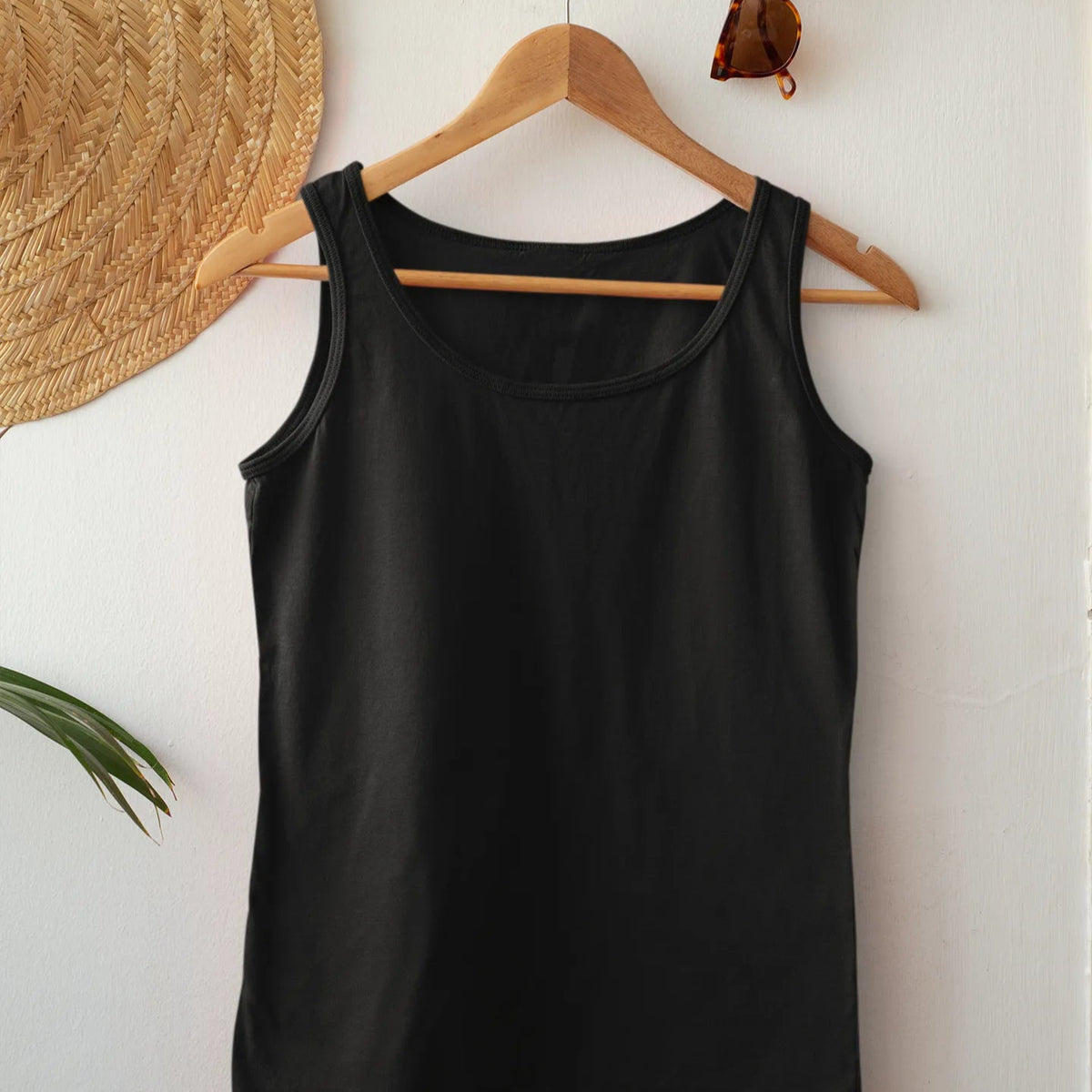 Black tank top on a wooden hanger against a white wall with a hat and sunglasses in the background.