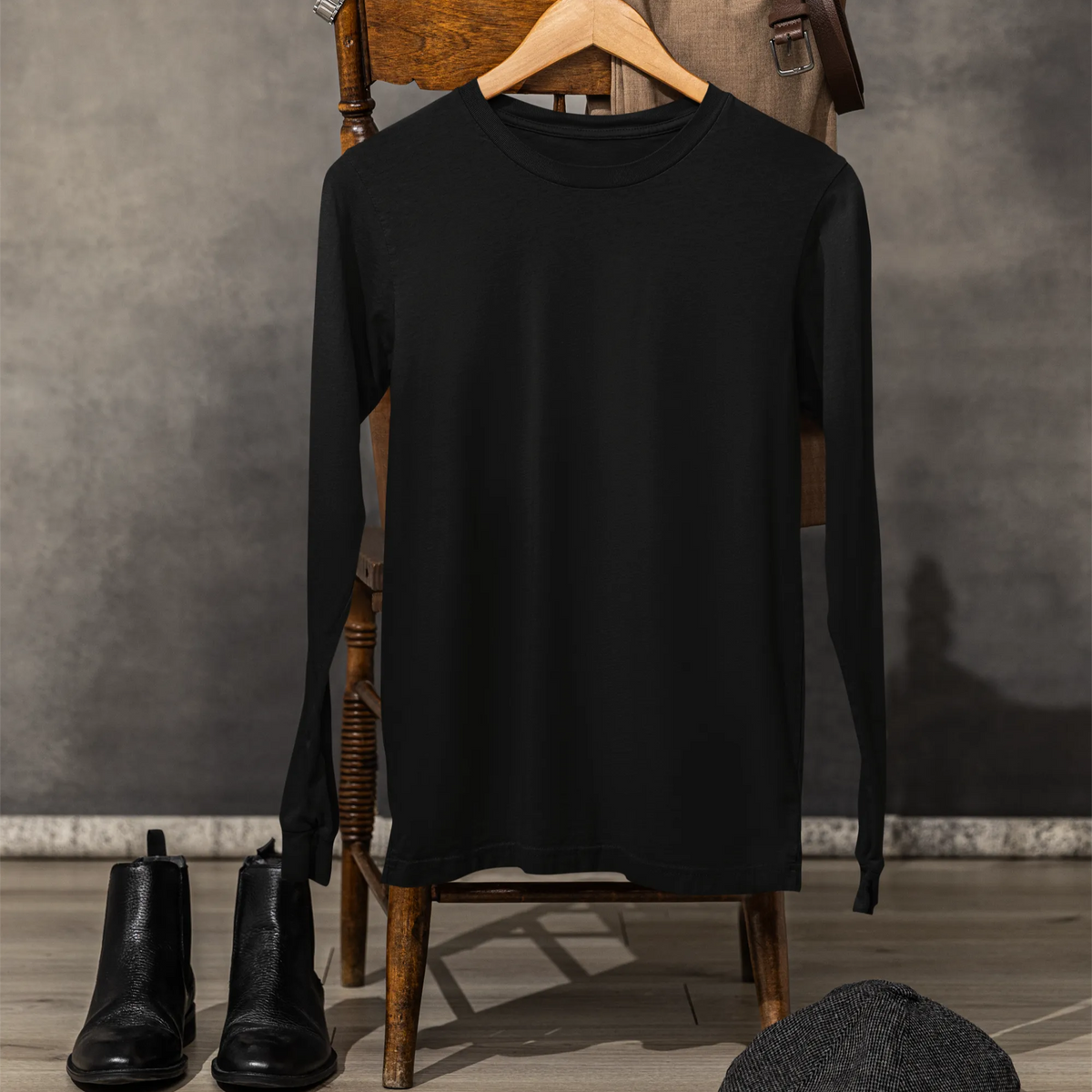 Black long-sleeve shirt on a wooden chair with black boots and a cap on the floor against a gray wall.