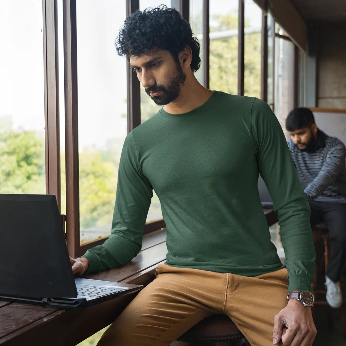 Man sitting at a table with a laptop, wearing a green shirt and beige pants, in a room with large windows.
