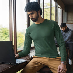 Man sitting at a table with a laptop, wearing a green shirt and beige pants, in a room with large windows.