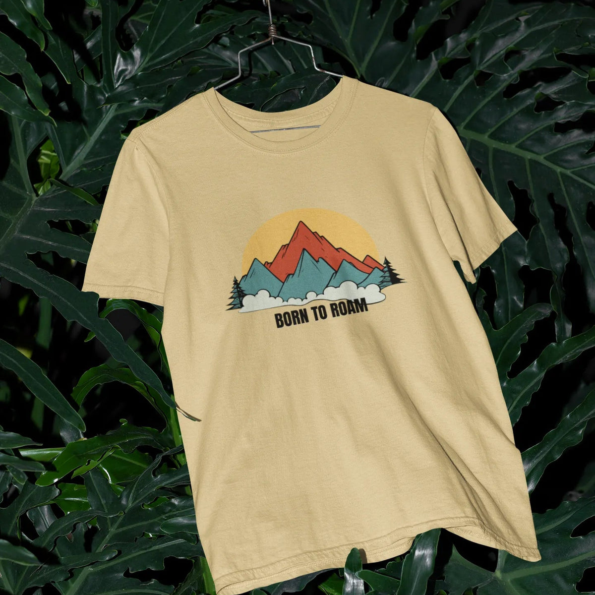 Beige t-shirt with mountain graphic and 'Born to Romm' text on a hanger against a leafy background