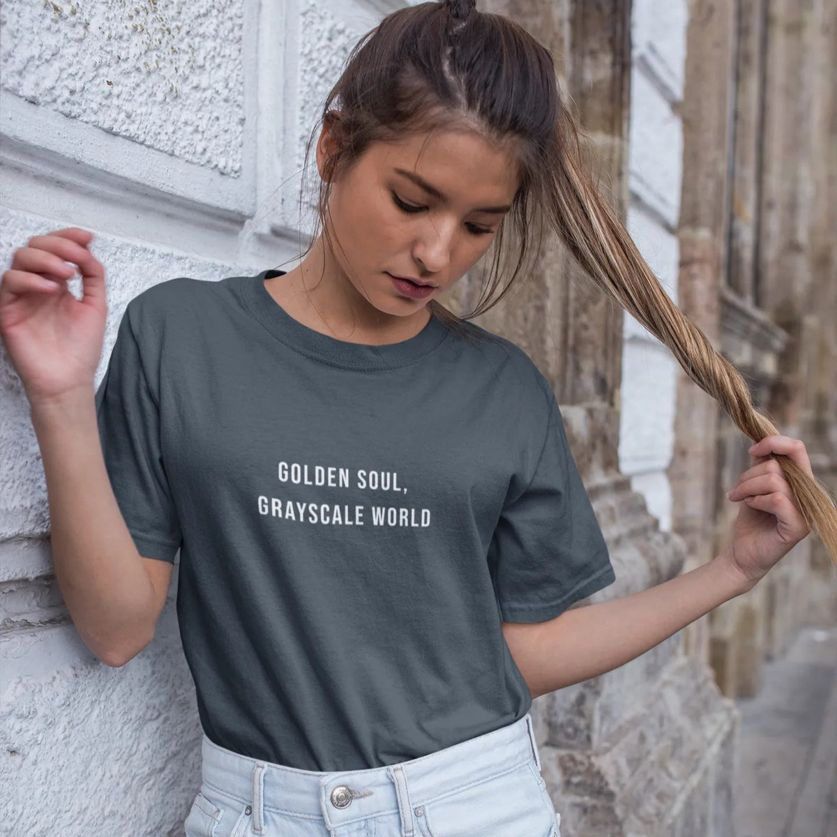 Woman wearing a dark gray t-shirt with 'Golden Soul, Grayscale World' text, standing against a stone wall.