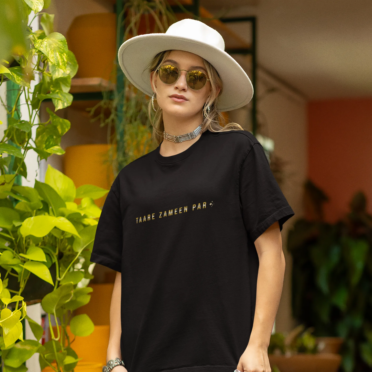 Person wearing a black t-shirt with text, white hat, and sunglasses in an outdoor setting with plants.
