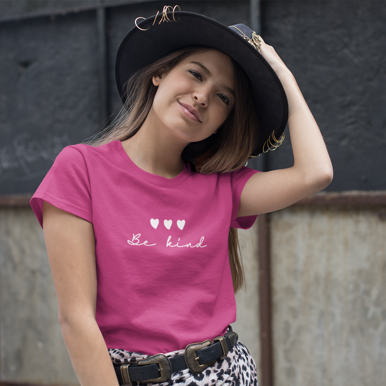Young girl wearing a pink t-shirt with 'Be kind' text and hearts, posing against a textured wall.