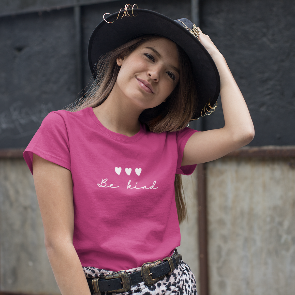 Young girl wearing a pink t-shirt with 'Be kind' text and hearts, posing against a textured wall.