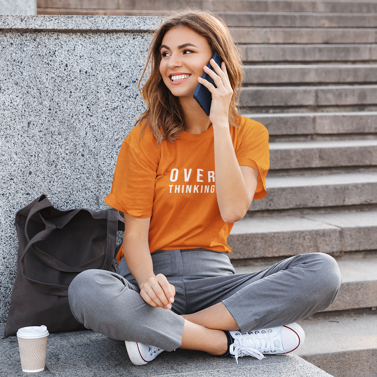 Woman sitting on steps talking on a phone with an 'Over Thinking' shirt.