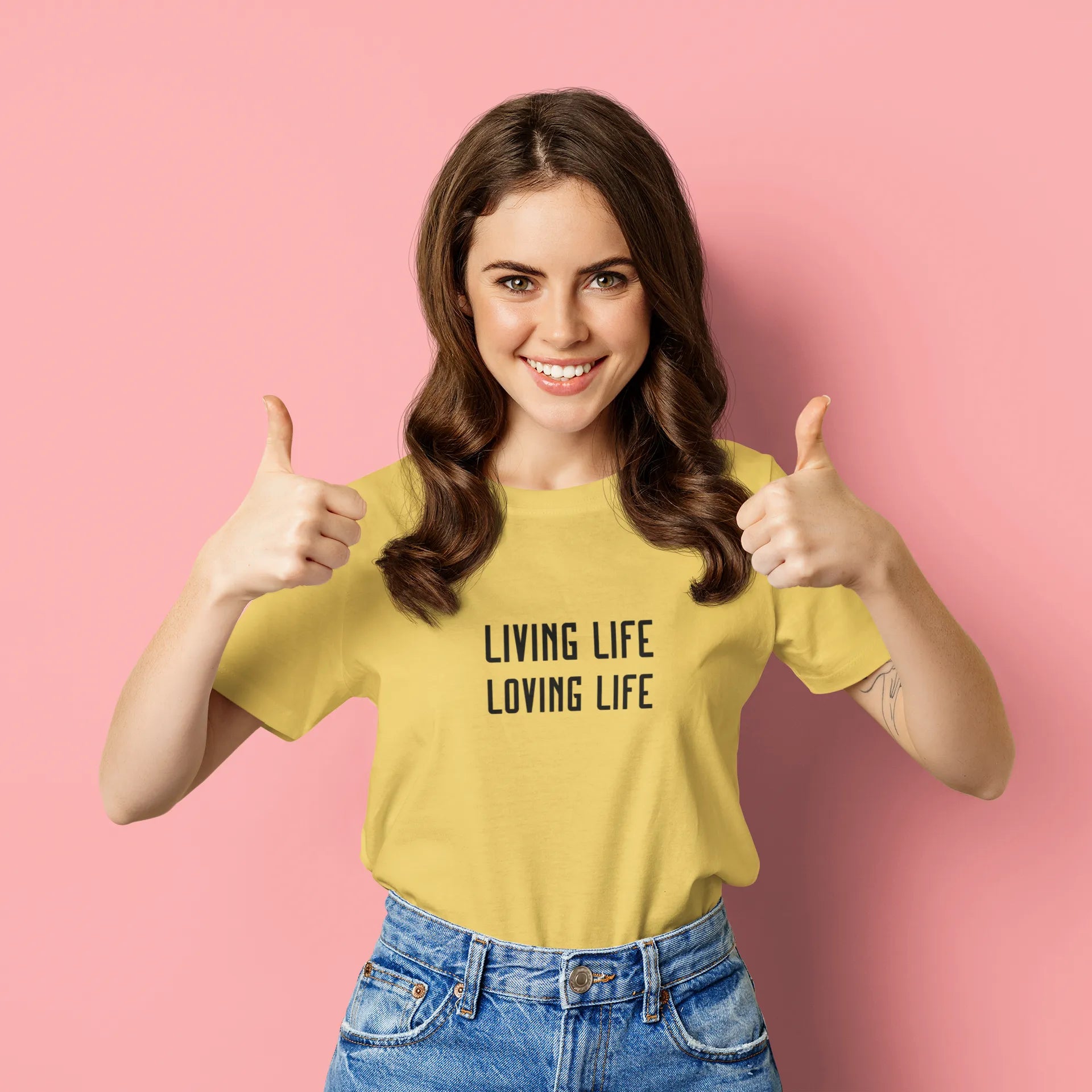 Woman wearing a yellow t-shirt with 'LIVING LIFE LOVING LIFE' text, giving thumbs up on a pink background