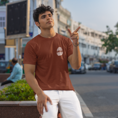Man in a brown t-shirt and white pants standing on a street with buildings and cars in the background.