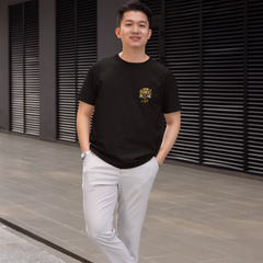 Man wearing a black t-shirt with a logo, light gray pants, and white sneakers standing on a pavement.