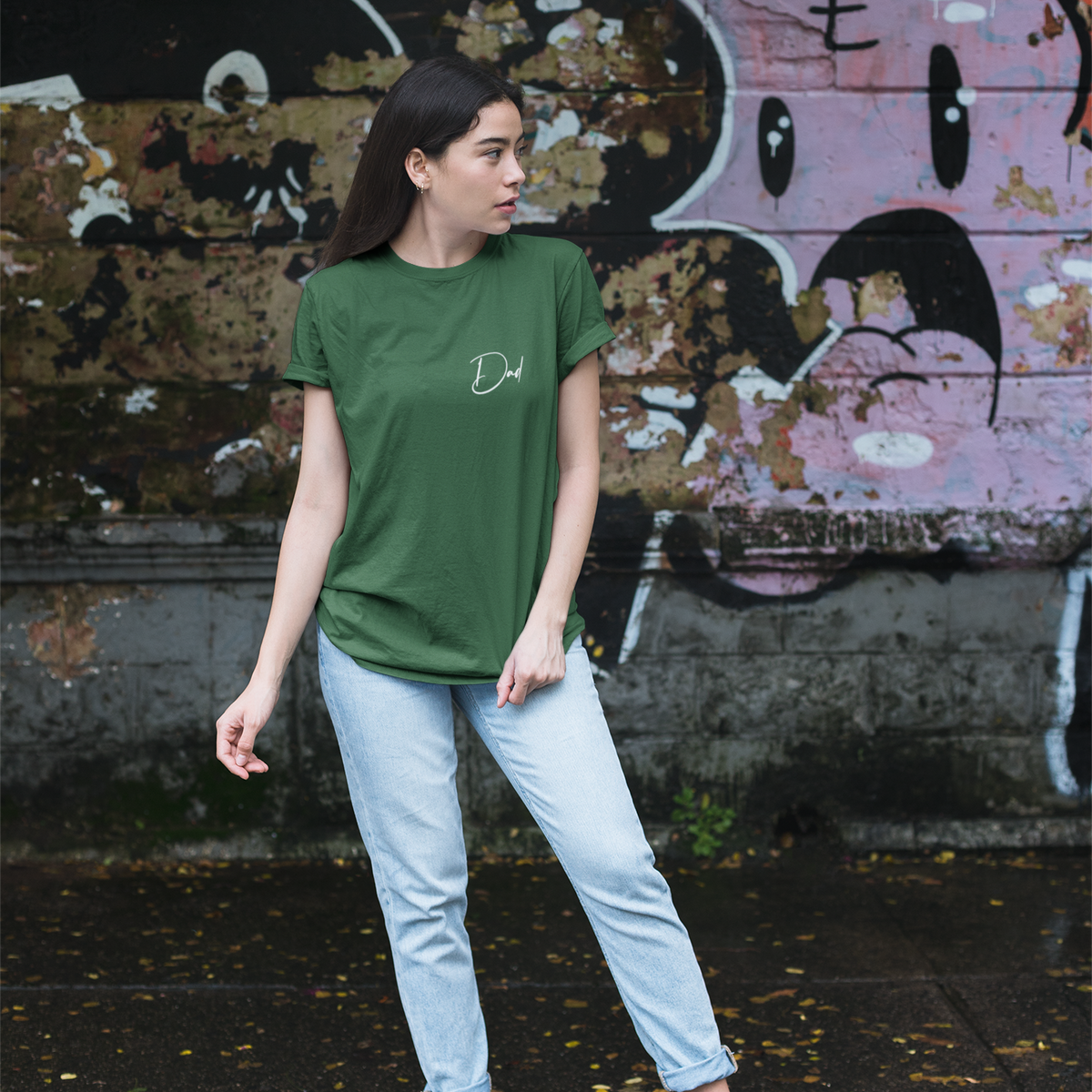 Person wearing a green t-shirt and light blue jeans standing in front of a graffiti-covered wall.