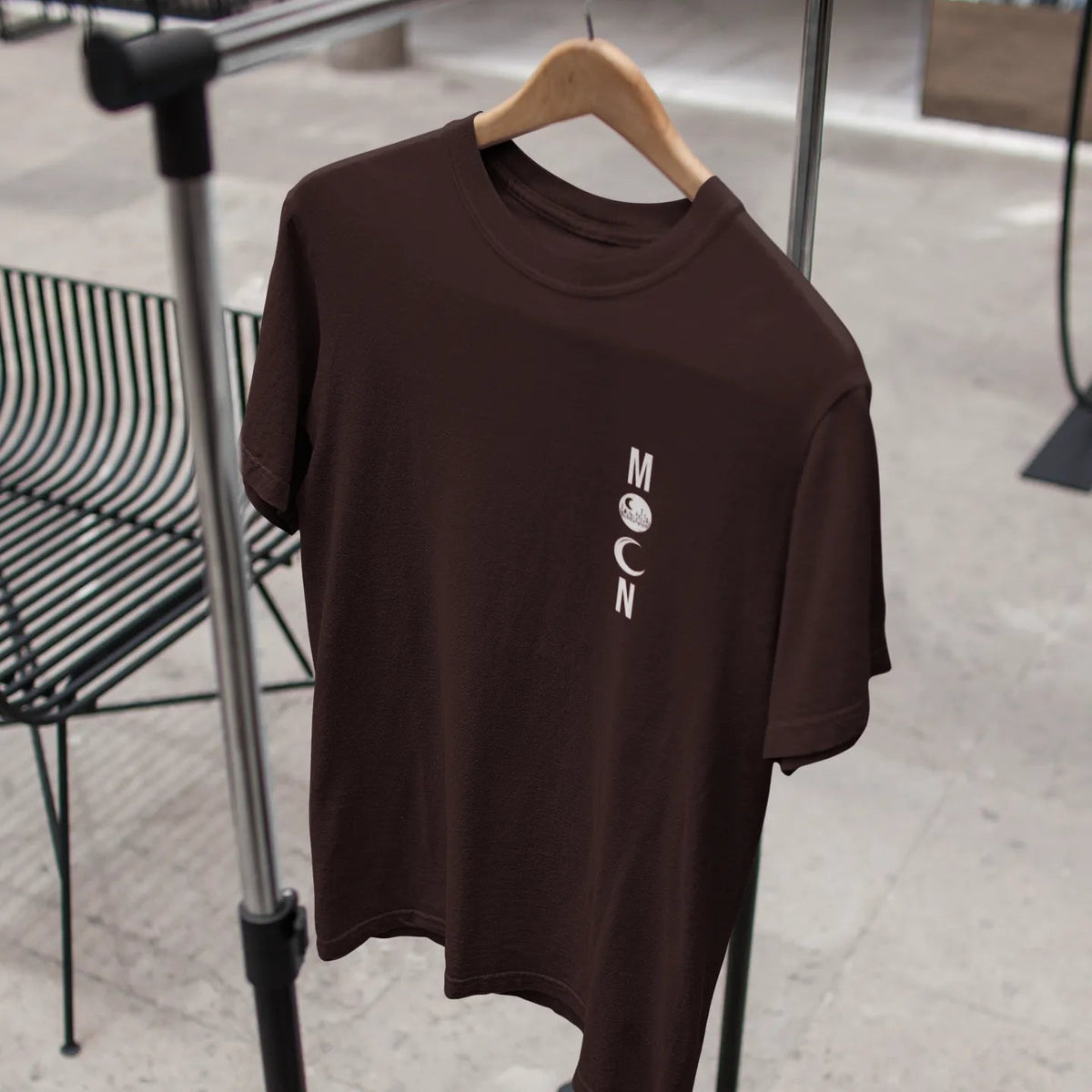 Brown t-shirt with 'MOON' logo on a hanger against a blurred indoor background