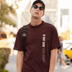 Man wearing a maroon t-shirt with text, sunglasses, and a cap in an urban setting.