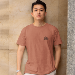 Man wearing a brown t-shirt with text standing against a textured wall.
