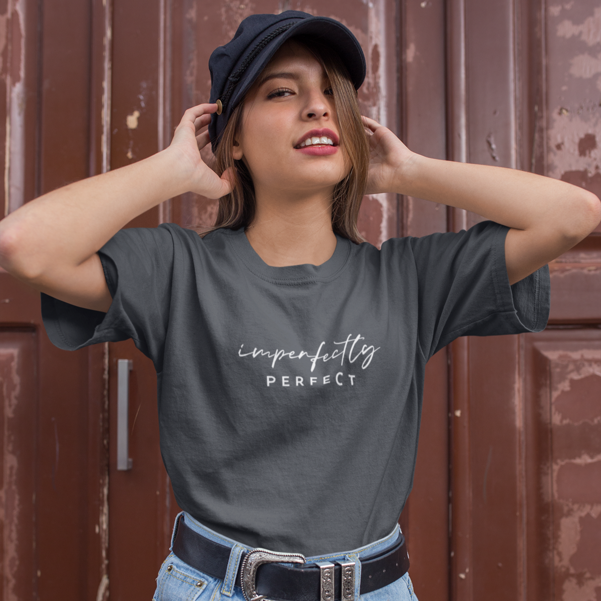 Woman wearing a gray t-shirt with 'imperfectly perfect' text, blue shorts, and a black cap against a brown background.