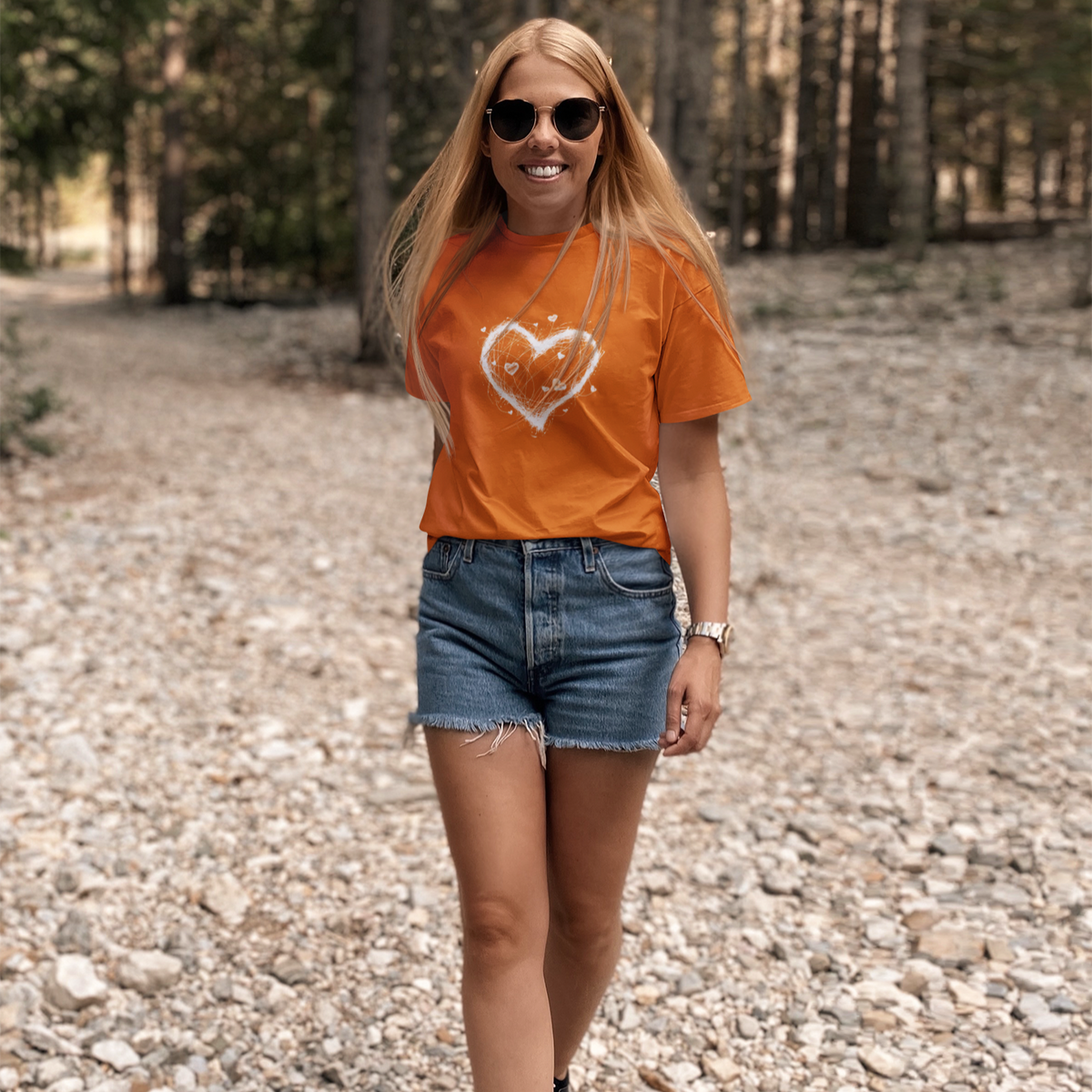 Woman wearing an orange t-shirt with a heart design and denim shorts walking on a gravel path.