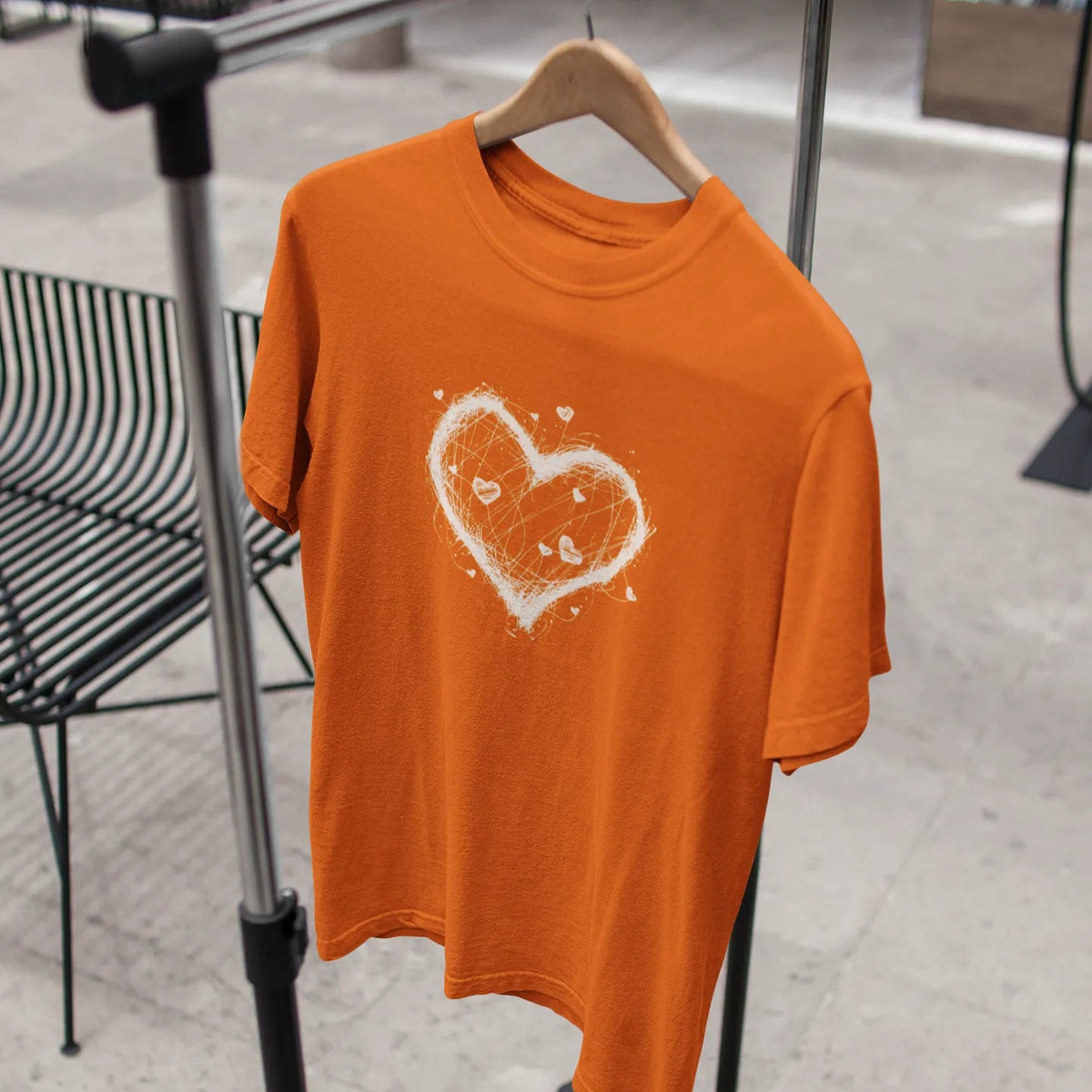 Orange t-shirt with a white heart design on a hanger against a blurred indoor background