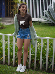 Woman wearing a brown t-shirt with a graphic design, blue denim shorts, and white sneakers, standing in front of a white fence.