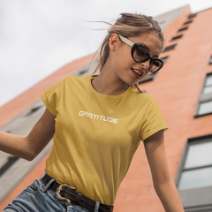 Woman wearing a yellow t-shirt with 'GRATITUDE' text, standing in front of a building.