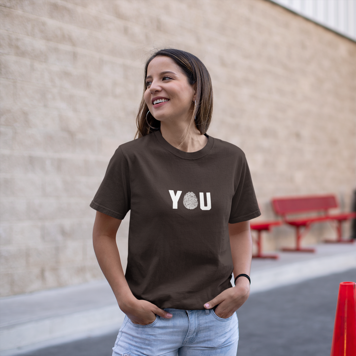 Woman wearing a brown t-shirt with 'YOU' printed on it, standing outdoors.