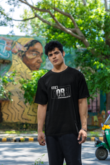 Man wearing a black t-shirt with text standing outdoors with greenery and a mural in the background