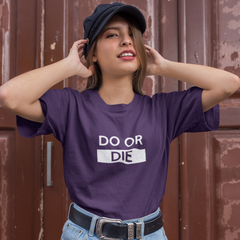 Person wearing a purple t-shirt with 'DO OR DIE' text, blue shorts, and a cap against a brown background.