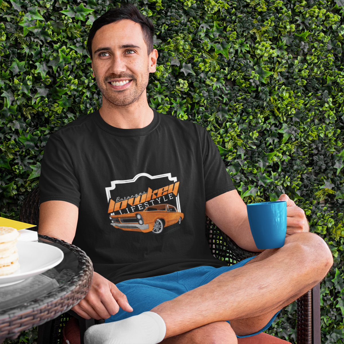 Man sitting outdoors with a plate of food and a mug, wearing a black t-shirt with a graphic design.