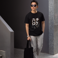 Man wearing a black t-shirt with white text and graphics, holding a black bag, standing against a gray wall.