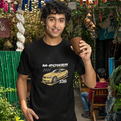 Man wearing a black t-shirt with a car graphic and text, holding a cup in an outdoor market setting.