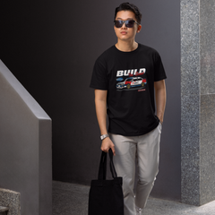 Man wearing a black t-shirt with graphics and text, holding a black bag, standing against a gray wall.