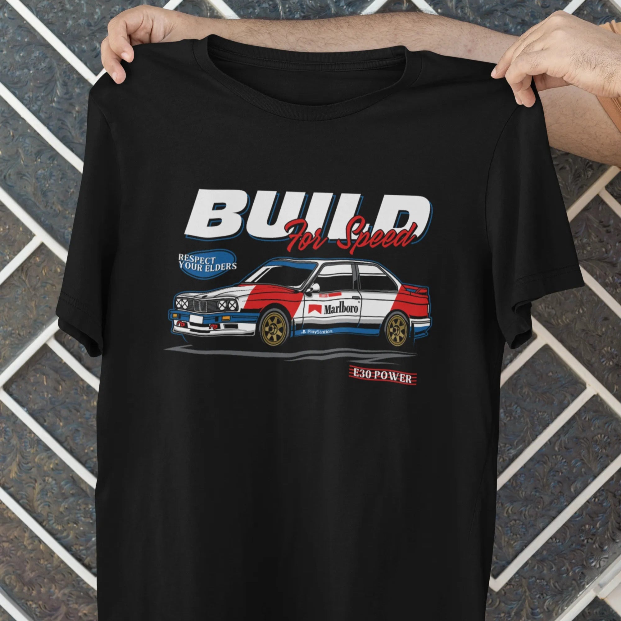Black t-shirt with a car graphic and text held by a person against a tiled wall.