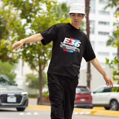 Person wearing a black t-shirt with a logo and text, standing on a street with cars and trees in the background.