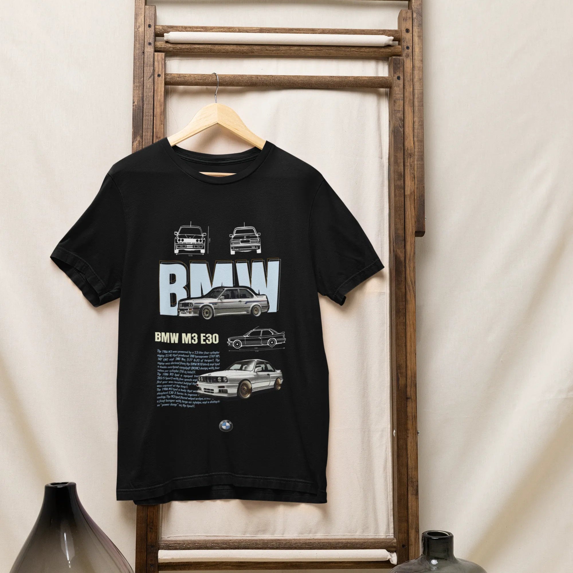 Black t-shirt with BMW logo and car illustrations on a wooden rack against a white curtain background.