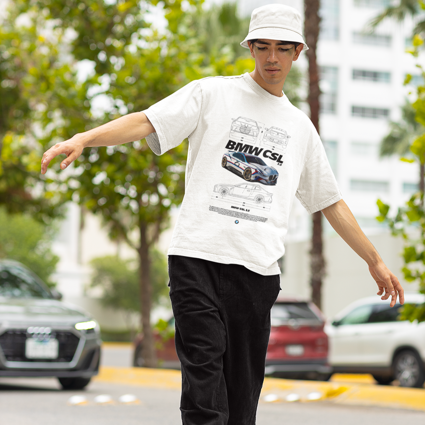 Person wearing a white t-shirt with a graphic design, black pants, and a white cap, standing on a street.