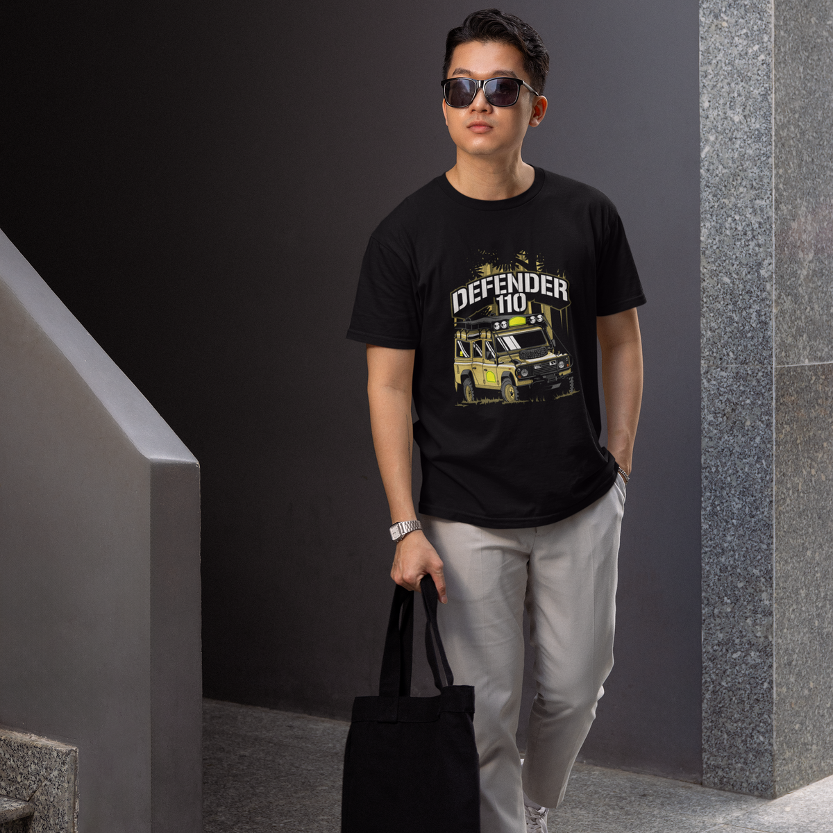 Man wearing a black t-shirt with graphic design, sunglasses, and holding a black bag in an indoor setting.