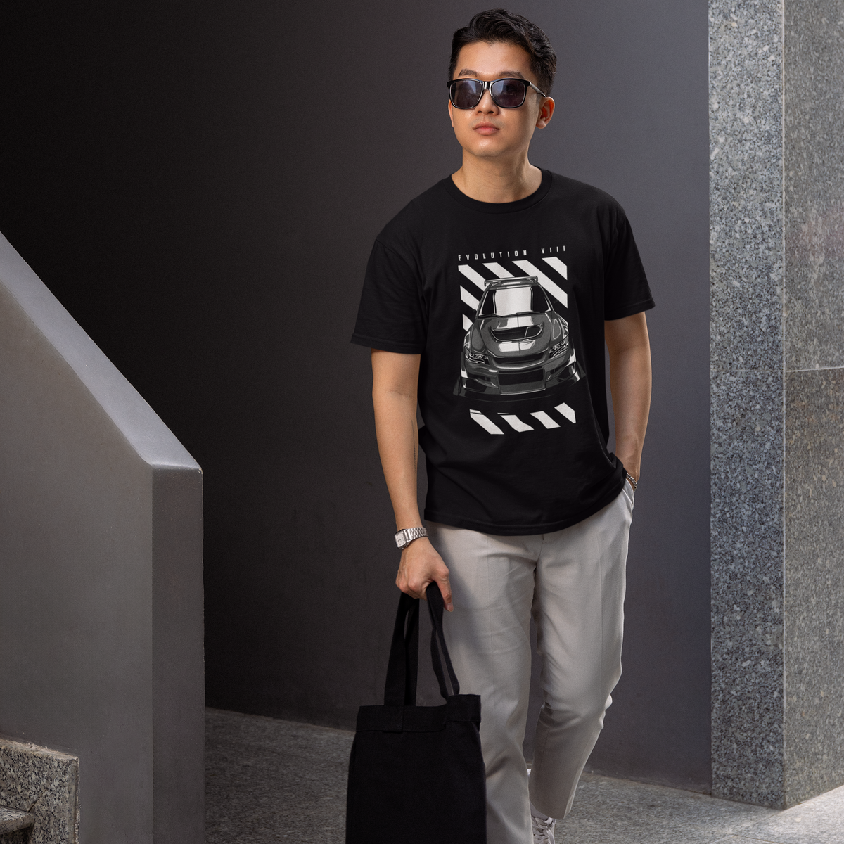 Man wearing a black t-shirt with a graphic design, holding a black bag, and standing in an indoor setting.