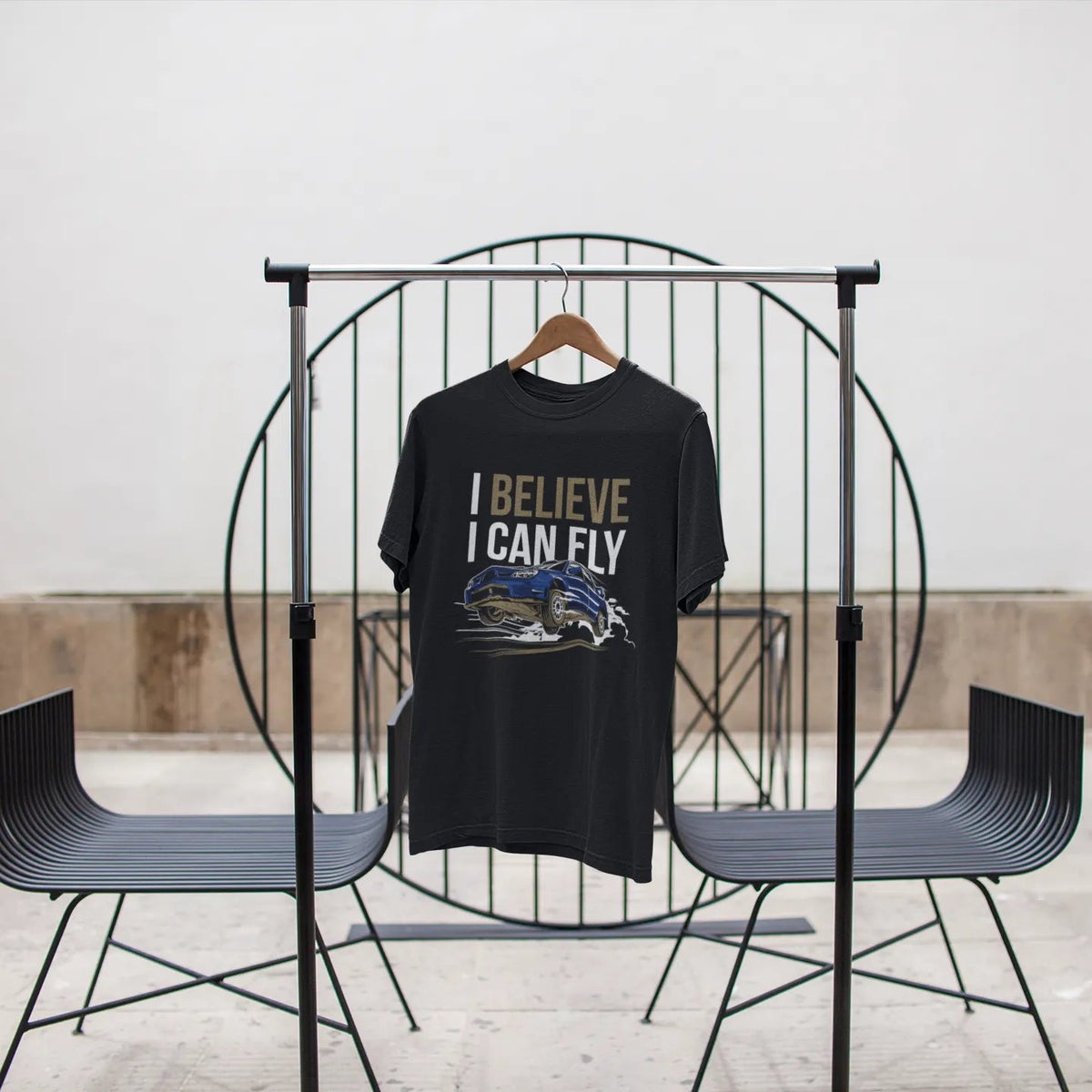 Black t-shirt with 'I Believe I Can Fly' text on a black metal stand against a white wall.