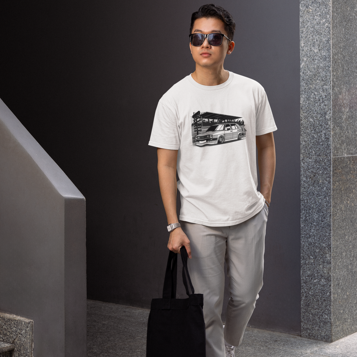 Man wearing sunglasses and a white t-shirt with a graphic design, holding a black tote bag, standing against a gray wall.