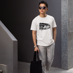 Man wearing sunglasses and a white t-shirt with a graphic design, holding a black tote bag, standing against a gray wall.