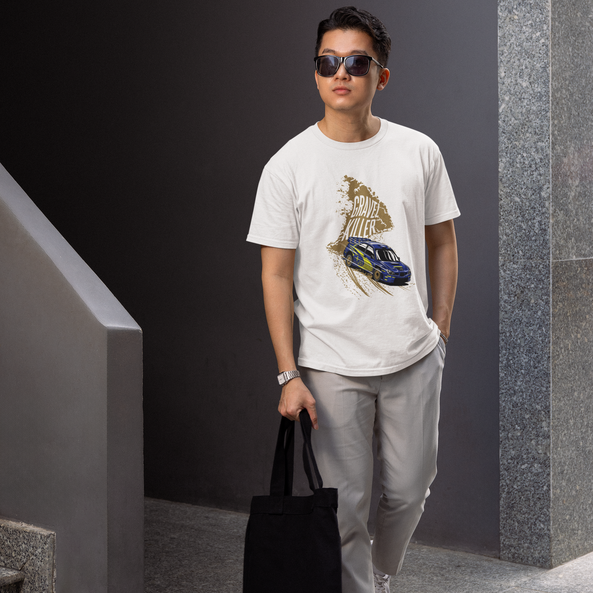 Man wearing a white t-shirt with a graphic design, holding a black bag, and standing in an indoor setting.