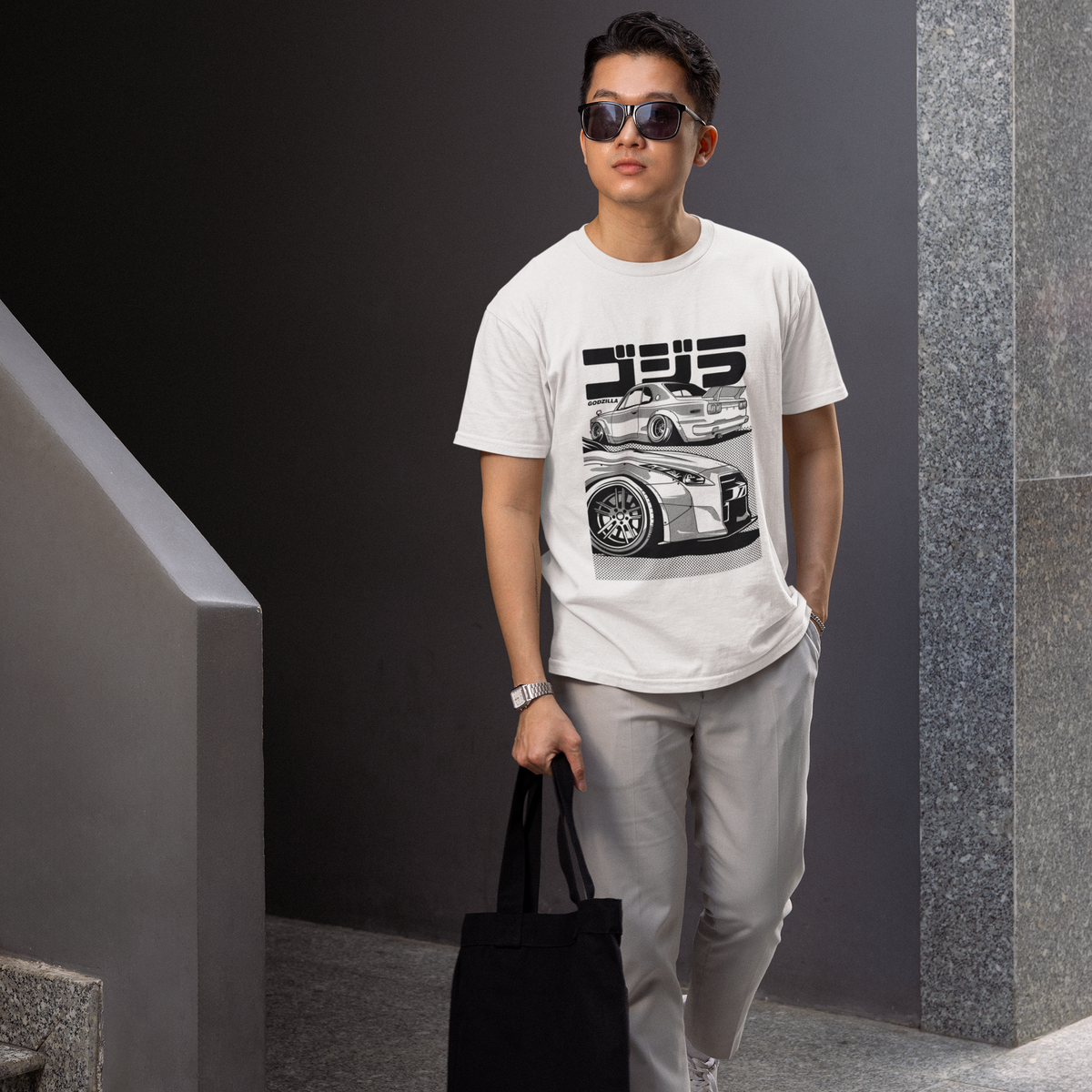 Man wearing a white t-shirt with a graphic design, holding a black bag, standing against a gray wall.