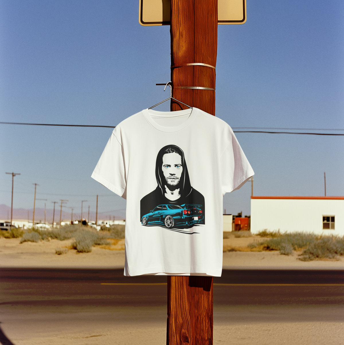 White t-shirt with a graphic design of a man and car hanging on a wooden pole in a desert setting.