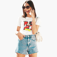 Woman wearing a white t-shirt with a graphic design and denim shorts, smiling and holding a phone.