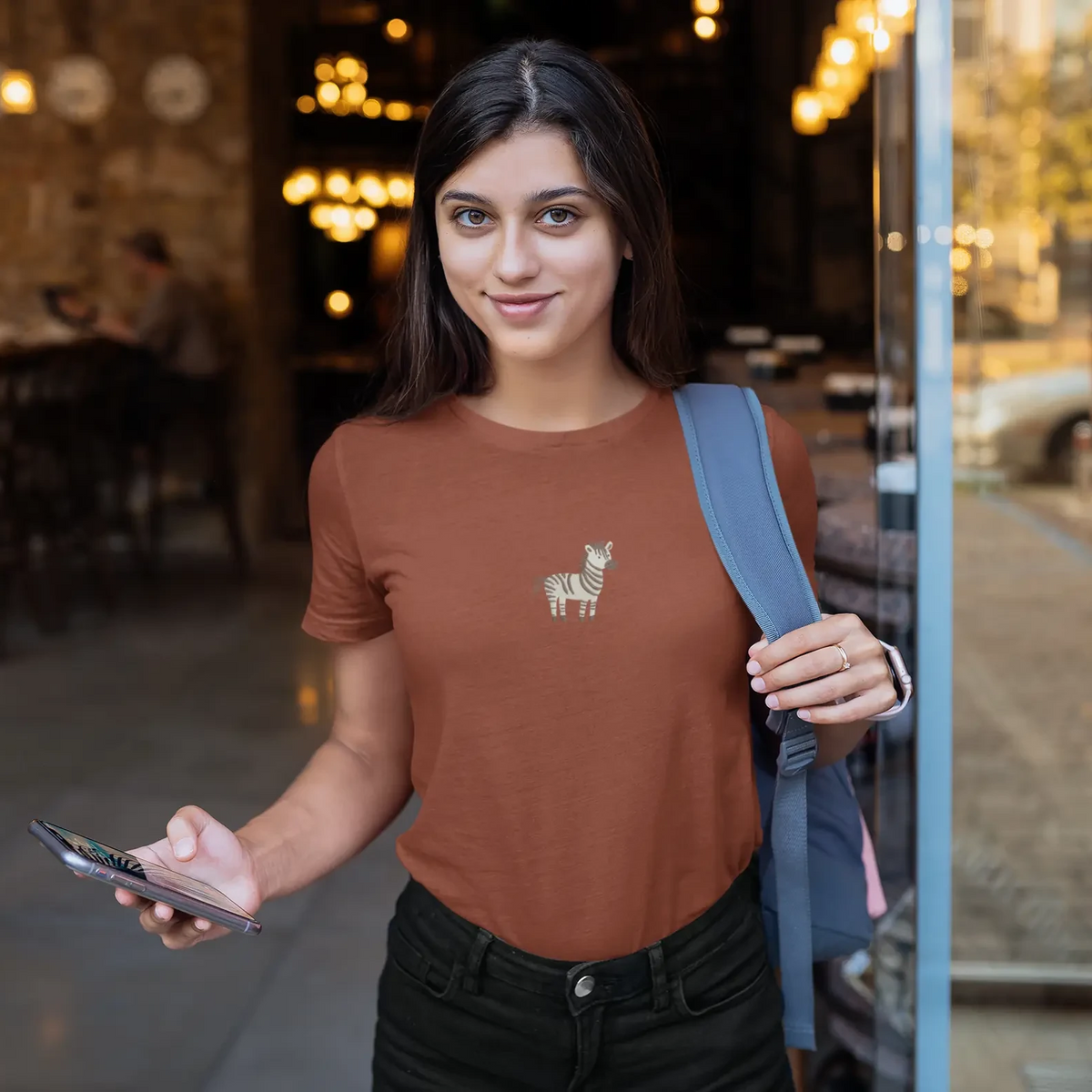 Woman wearing a brown t-shirt with a llama design, holding a phone, standing in a casual setting.