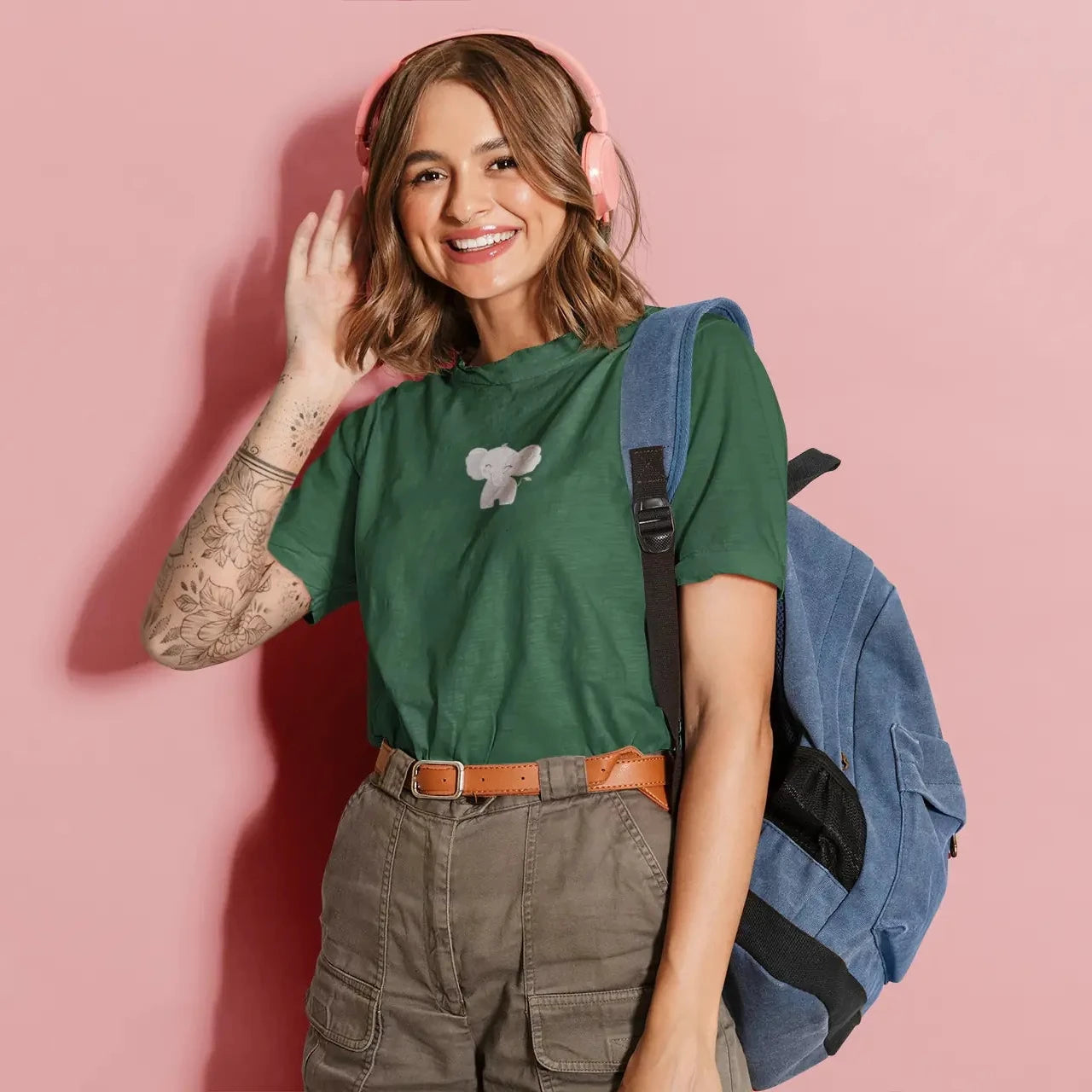 Woman wearing a green t-shirt with a small elephant design, holding a blue backpack against a pink background