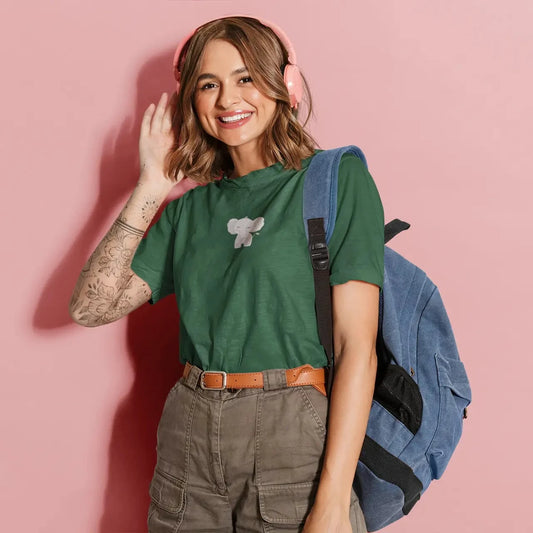 Woman wearing a green t-shirt with a small elephant design, holding a blue backpack against a pink background