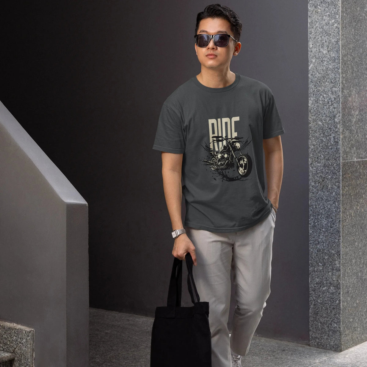 Man wearing sunglasses and a dark t-shirt with a graphic design, holding a black tote bag, standing against a gray wall.