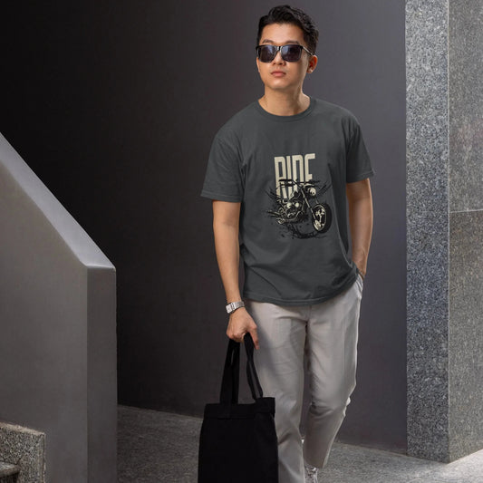 Man wearing sunglasses and a dark t-shirt with a graphic design, holding a black tote bag, standing against a gray wall.